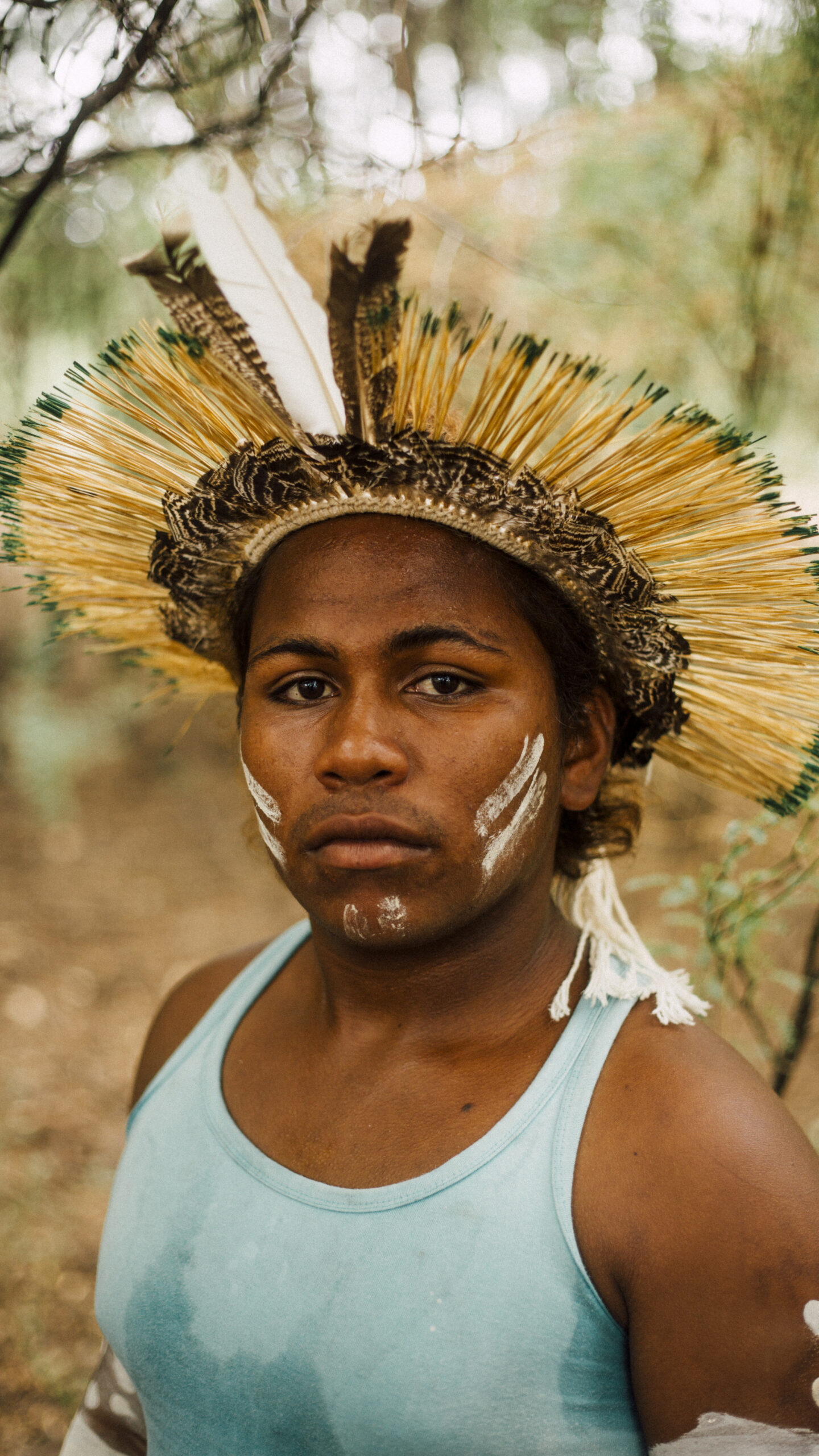 Portrait from the 'Origem' series, featuring illustrative background added through AR technology. A person wearing a blue vest with a headdress made of natural materials. They have two white stripes of paint across each cheek and on the chin. The background is blurred and shows a natural landscape with a tree closely behind in the top left corner.