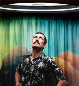 A portrait of artist Ivan Morison standing in front of colourful curtains looking up towards a skylight