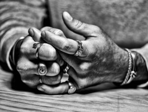 A black and white close up of some hands on a board, those of Eduardo Gonzalez. His hands are resting on their backs and their hands are intertwined. Eduardo has a variety of eye-catching rings, including a nail on his index finger and an eye on his right middle finger.